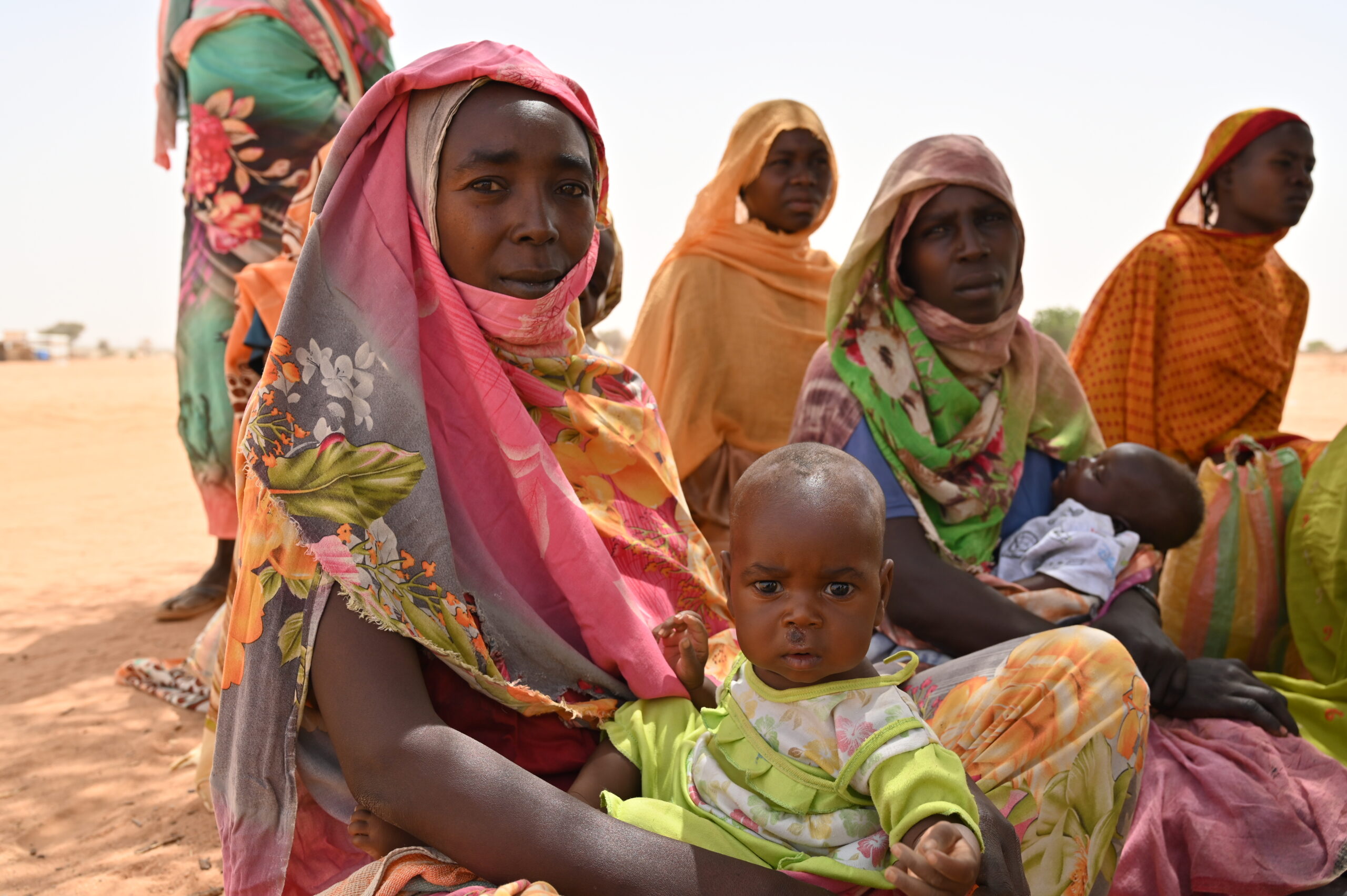 A group of women dressed in colourful clothes and wearing headscarves are sitting on sand. Two of the women are holding babies.