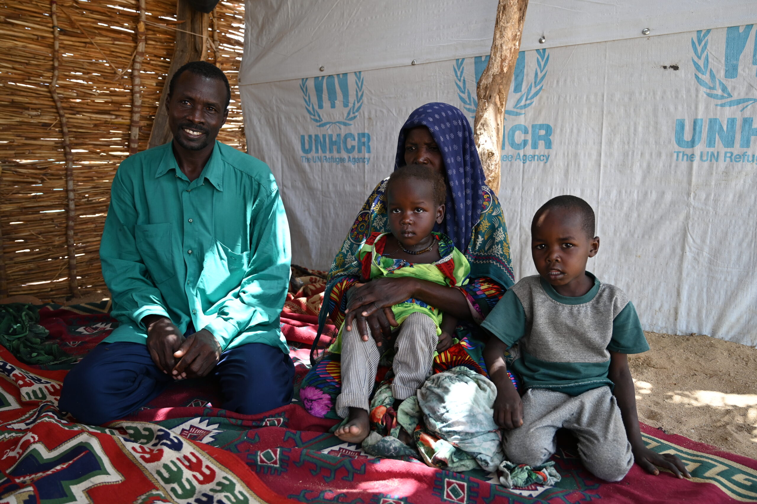 A family sits on their knees inside a tent where the walls display the UNHCR logo. The sand ground is padded with a colourful and patterned blanket in which they sit upon. A man smiles on the far left, a woman looks down while holding a child, the child being held gazes at the camera and a child on the far left also looks at the camera.