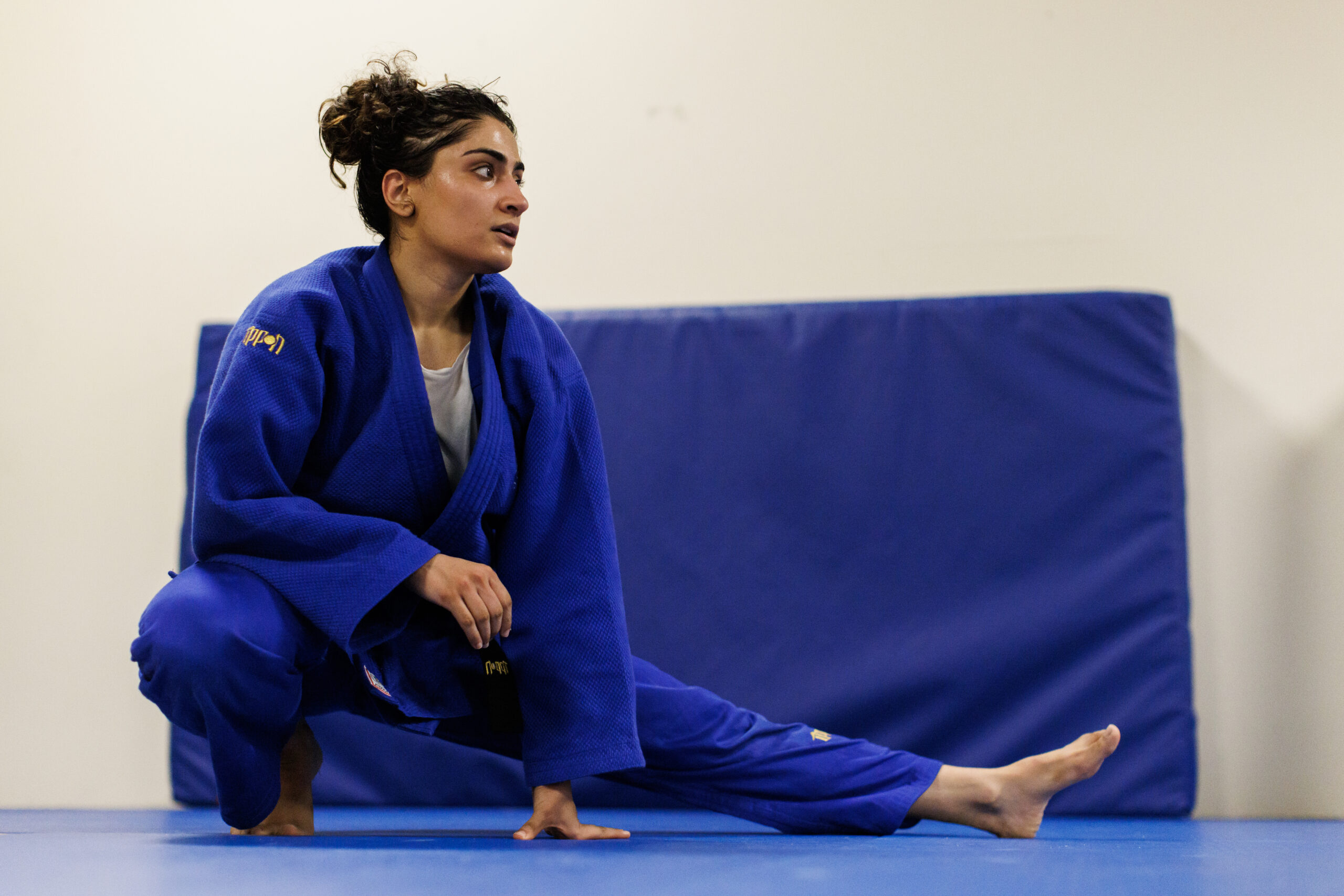 A female judo wrestler wearing a blue judo uniform does a "Cossack split" stretch on a wrestling mat, squatting on her right leg while her left leg is outstretched to the left side.